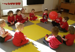 Widok na siedzące w kręgu dzieci, które w rytm muzyki podają sobie balonika.